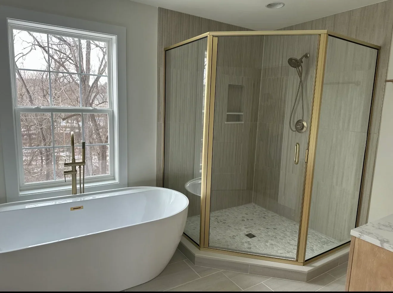 Freestanding white tub and frameless glass shower with brass trim, gray tile, pebble floor