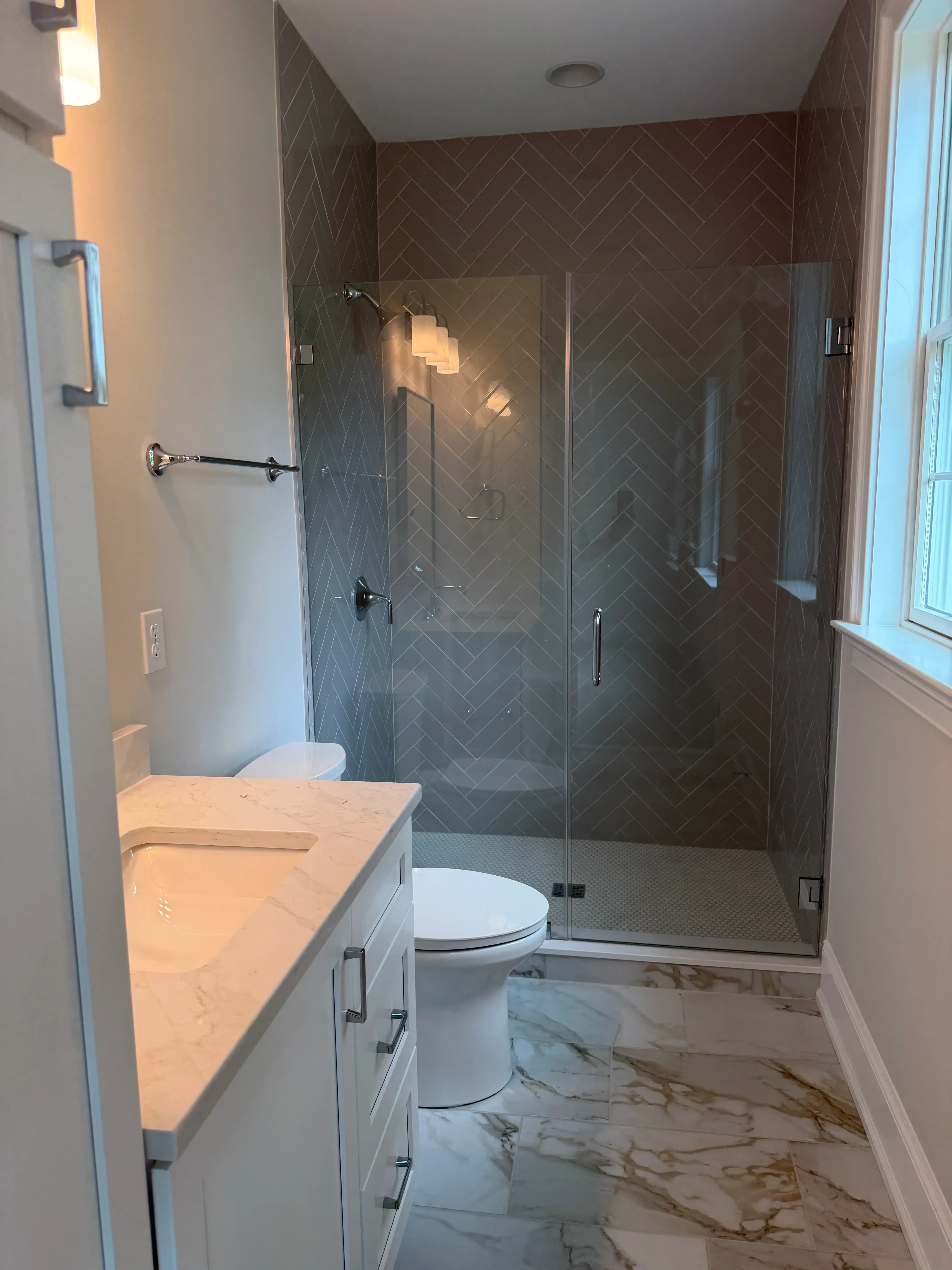 Walk-in shower with herringbone tile, frameless glass enclosure, and marble vanity